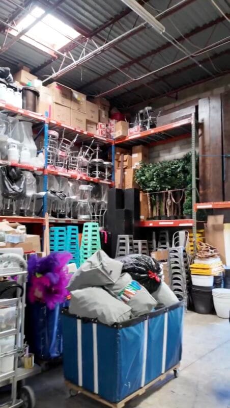 Event rental warehouse interior with shelving units stocked with chairs, glassware, and boxes, plus a rolling laundry cart loaded with linens and a purple feather decoration in the foreground