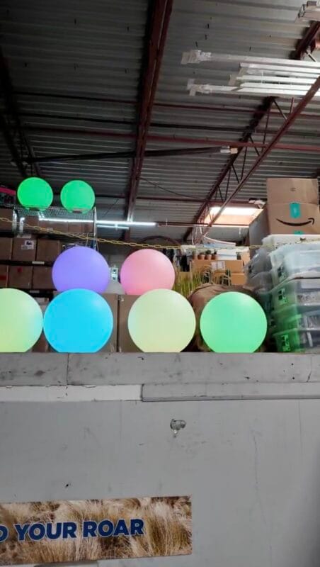 Row of illuminated globe light balls in green, purple, pink, blue, and teal displayed on a shelf inside an event rental warehouse