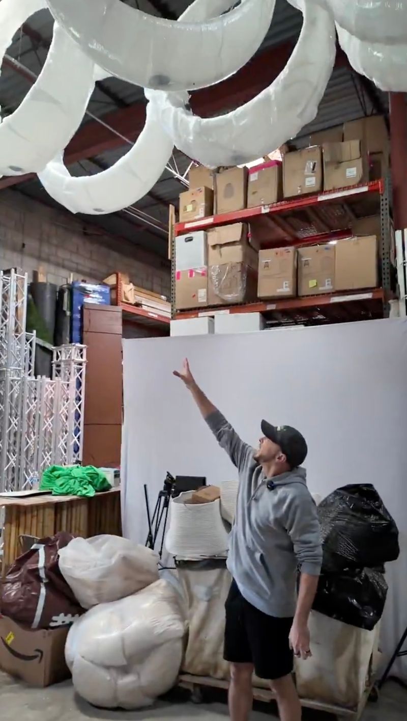 Jacob Becker pointing up at large white circular balloon decor elements hanging from the ceiling in an event decor warehouse stocked with shelves and props
