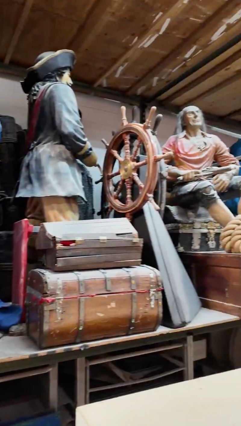Pirate-themed event decor props in a warehouse, including two life-size pirate mannequins, a ship's steering wheel, treasure chests, and an oar
